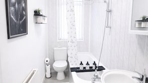 Een moderne badkamer met een wit toilet, badkuip en wastafel. Er is een douchekop, een handdoekhouder en een lijst met een foto aan de muur.