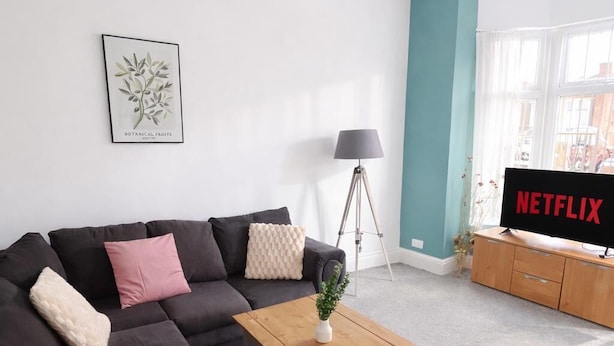 Een moderne woonkamer met een donkere bank, een houten salontafel, een vloerlamp en een tv waarop 'NETFLIX' staat.