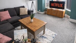 Een moderne woonkamer met een donkere bank, een houten salontafel met een welkomstbord, een koffiepot en twee kopjes. Een Netflix tv is aan de muur bevestigd, en er is een raam met jaloezieën.