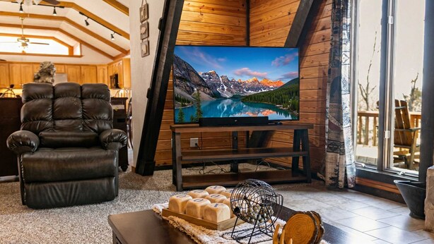 A cozy living room with a leather sofa, a wooden coffee table, and a flat-screen TV displaying a mountain landscape. - White River Escape with BBQ & Private Backyard (Norfork)