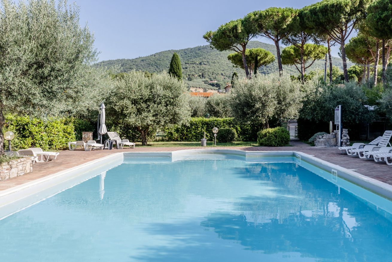 Ein Swimmingpool mit Liegestühlen, umgeben von Grünflächen und Bäumen, mit Blick auf Hügel im Hintergrund.