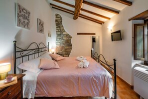 A bedroom with a bed, a nightstand, a lamp, a stone wall, a wooden ceiling beam, and a television. - GuestHost - Cozy 50m2 apartment equipped with all comforts, ideal for two people, located on the second floor of a renovated farmhouse (stairs to access it).The accommodation is located inside a former oil mill and offers its guests many shared services: (Passignano sul Trasimeno)