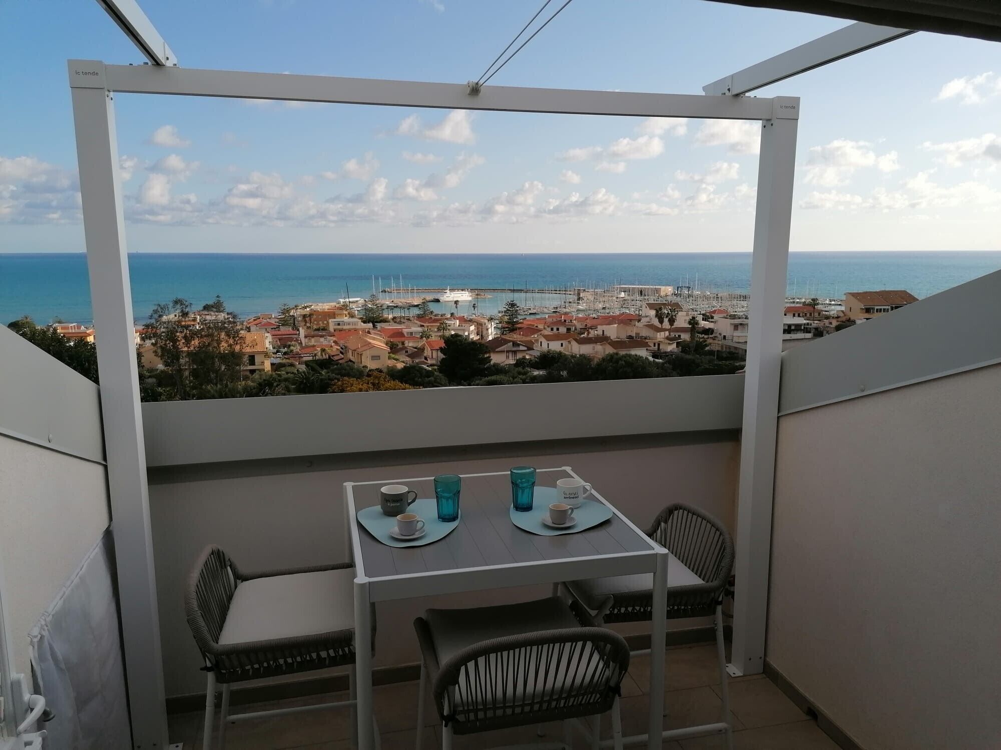 A balcony with a table and chairs, overlooking a coastal town with buildings and boats.