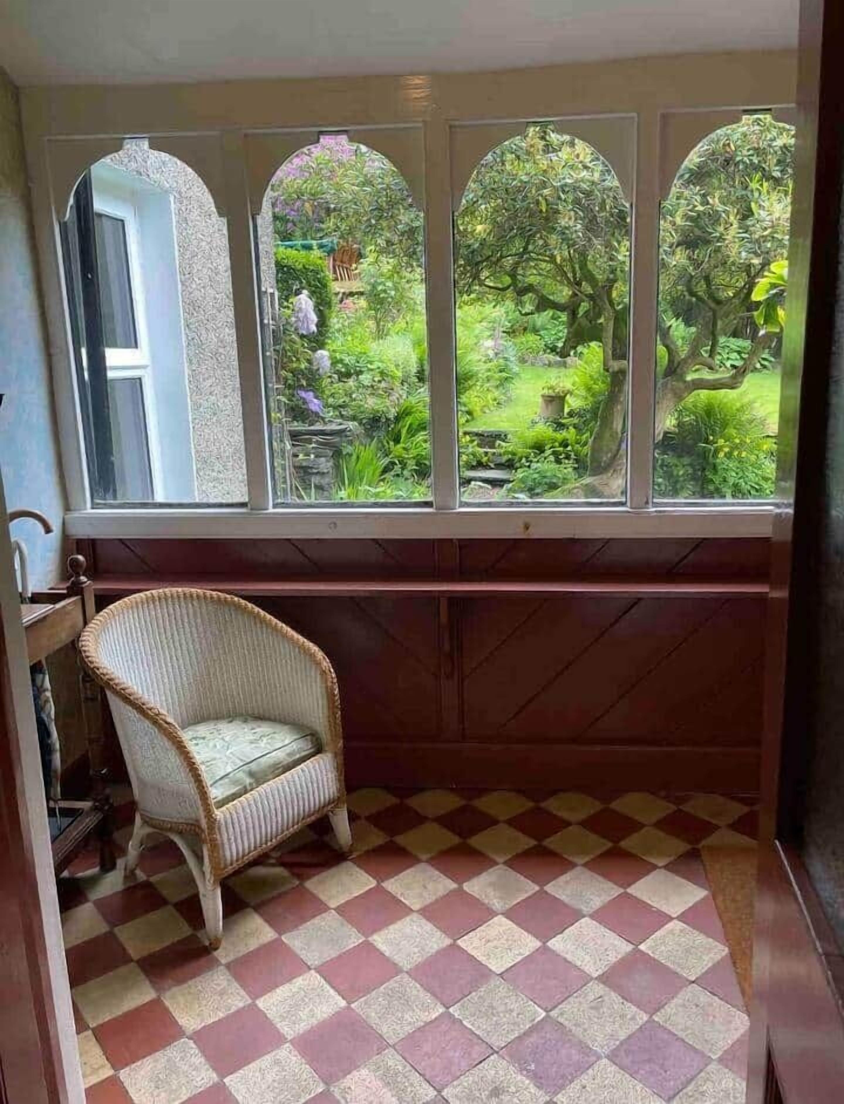 A room with a checkered floor, a wicker chair, and a window with a view of a garden.