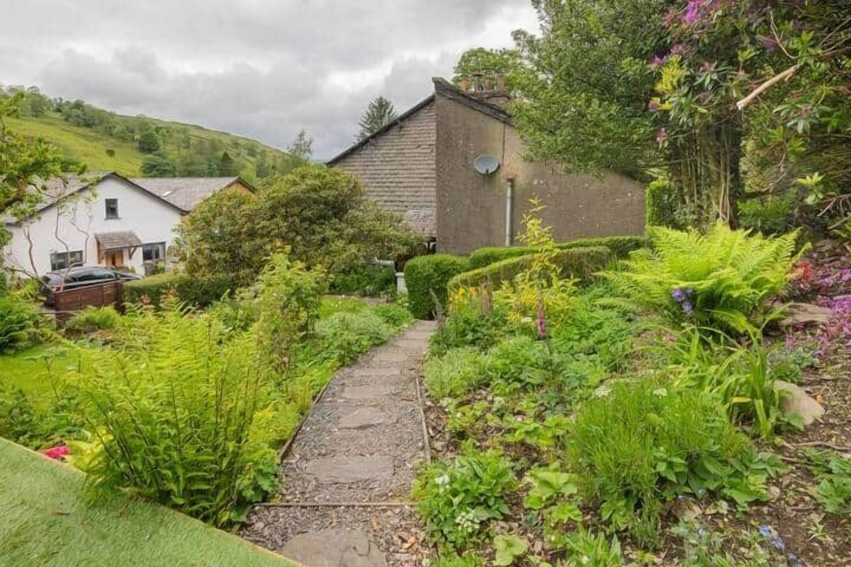 A garden pathway leading to a rustic building surrounded by lush greenery and flowering plants.