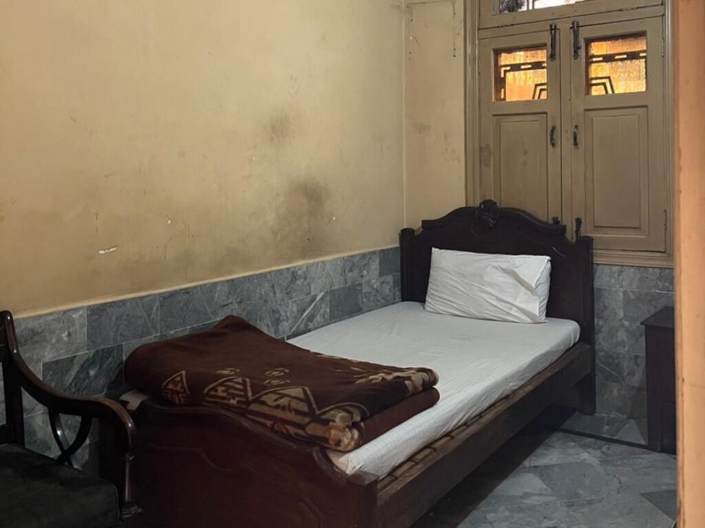 Una cama individual con una manta marrón, cabecera de madera y almohada blanca en una habitación con pared de piedra y armarios de madera.
