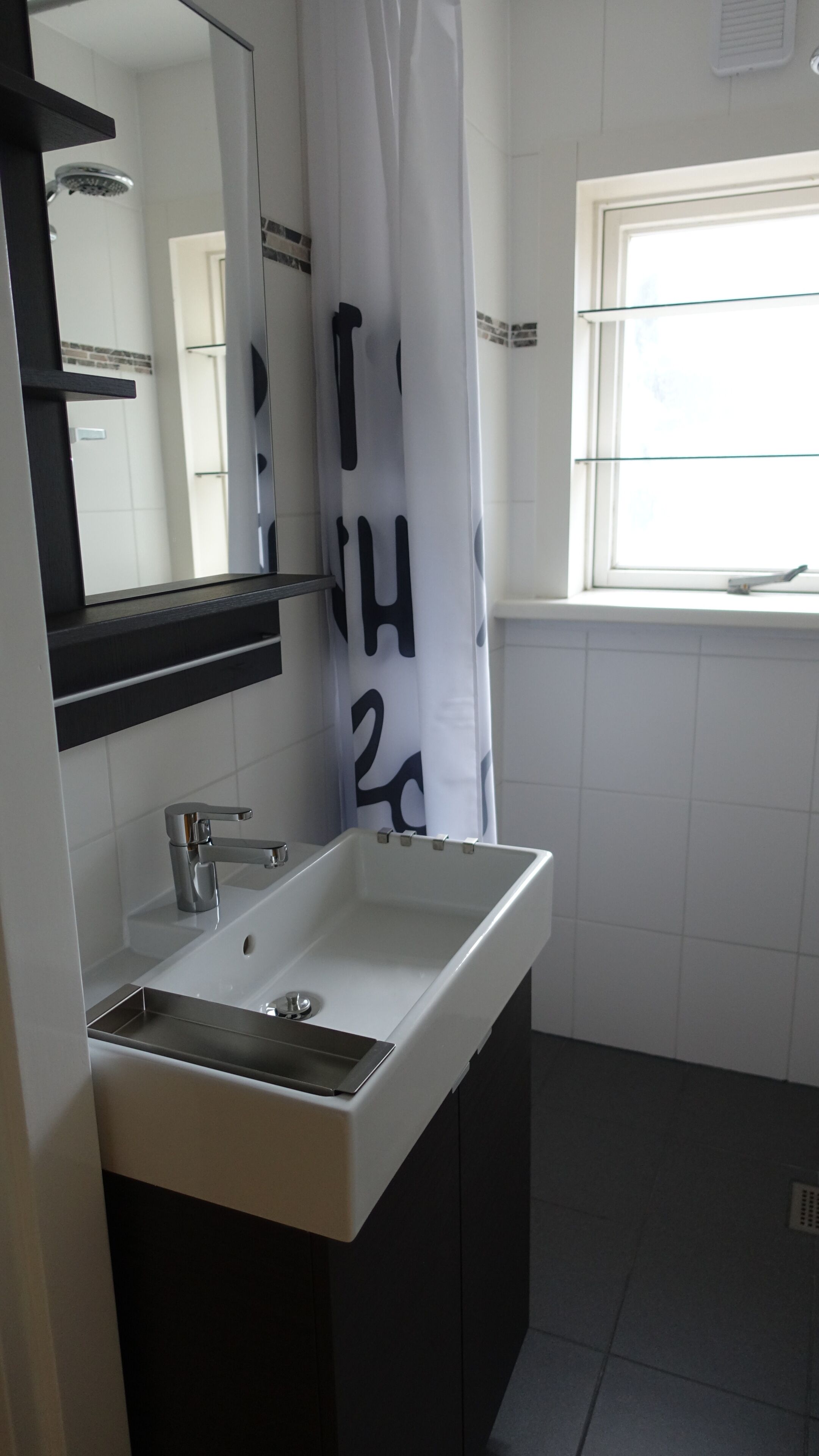 Een badkamer met een wit wastafel, een spiegel, een douche en een raam.