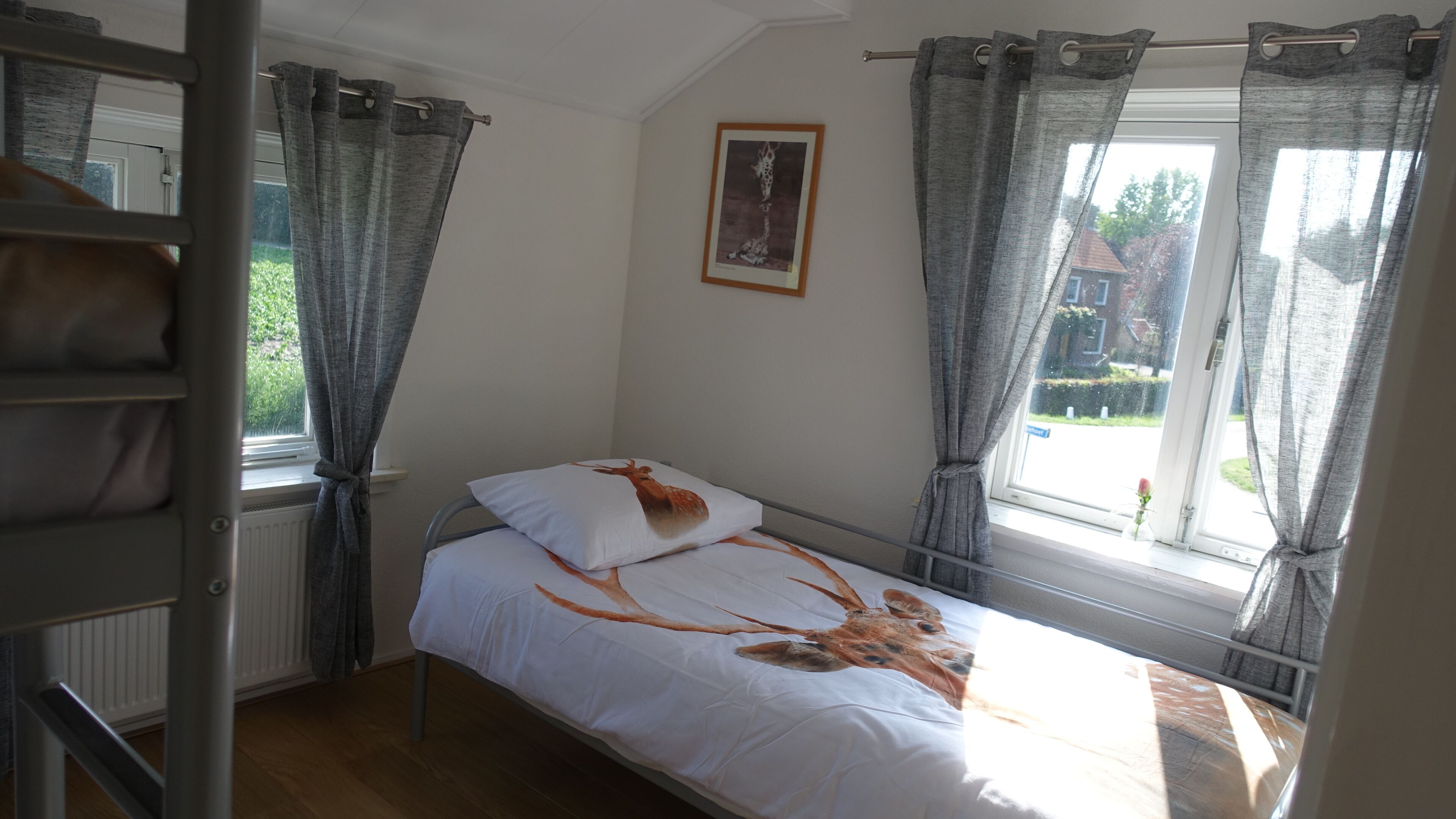 A single bed with a deer print pillow, a radiator, a window with curtains, and a framed picture on the wall.