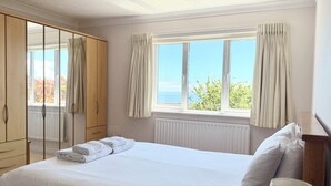 A bedroom with a large bed, a wardrobe, a window with curtains, and a view of the sea.