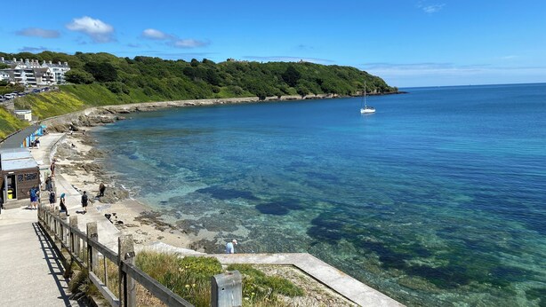 A coastal area with a clear view of the sea, a wooden walkway along the shore, and a sailboat in the distance.