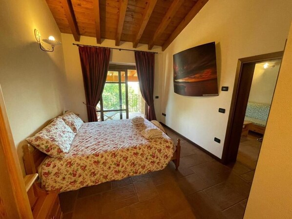 Un dormitori amb llit de fusta, una finestra gran amb cortines, un quadre penjat a la paret i una porta que dóna a una altra habitació.