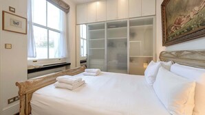 Una cama bien hecha con sábanas y toallas blancas, un amplio ventanal con cortinas, un armario con puertas de vidrio y un cuadro en la pared.