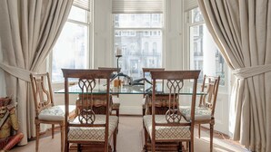 Una habitación con cuatro sillas de madera con asientos acolchados, una mesa con superficie de vidrio y vistas a un edificio a través de un ventanal con cortinas.