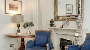 Una habitación con dos sillones azules, una mesa redonda de madera con una lámpara, una chimenea y un espejo con un marco decorativo.