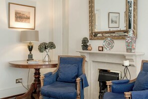 A room with two blue armchairs, a round wooden table with a lamp, a fireplace, and a mirror with a decorative frame. - Charming 2 bedroom flat - Knightsbridge (Greater London)