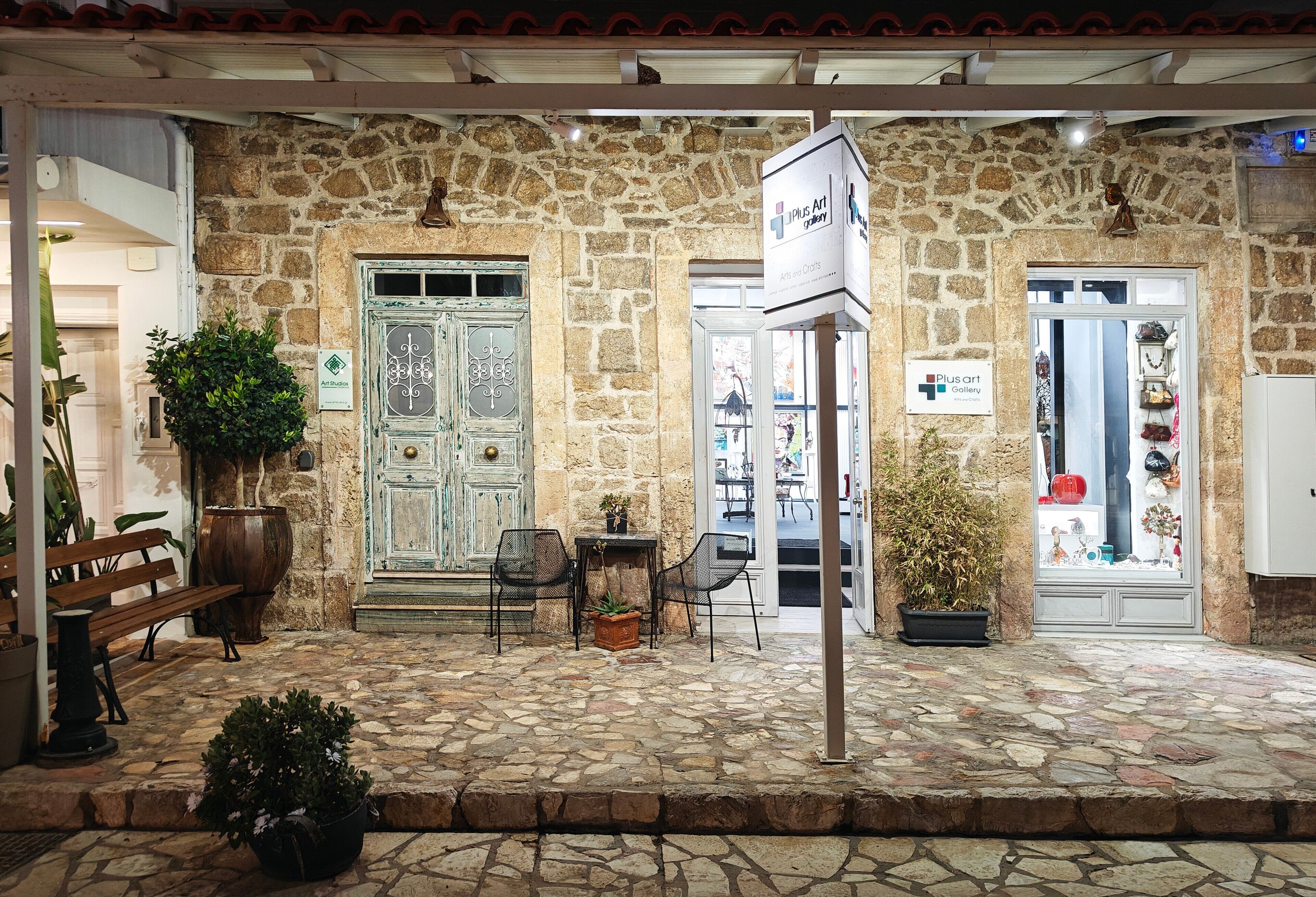 A stone building with a sign for 'Plus Art Gallery' and a door labeled 'All Rooms.' There are outdoor seating areas with chairs and a bench.