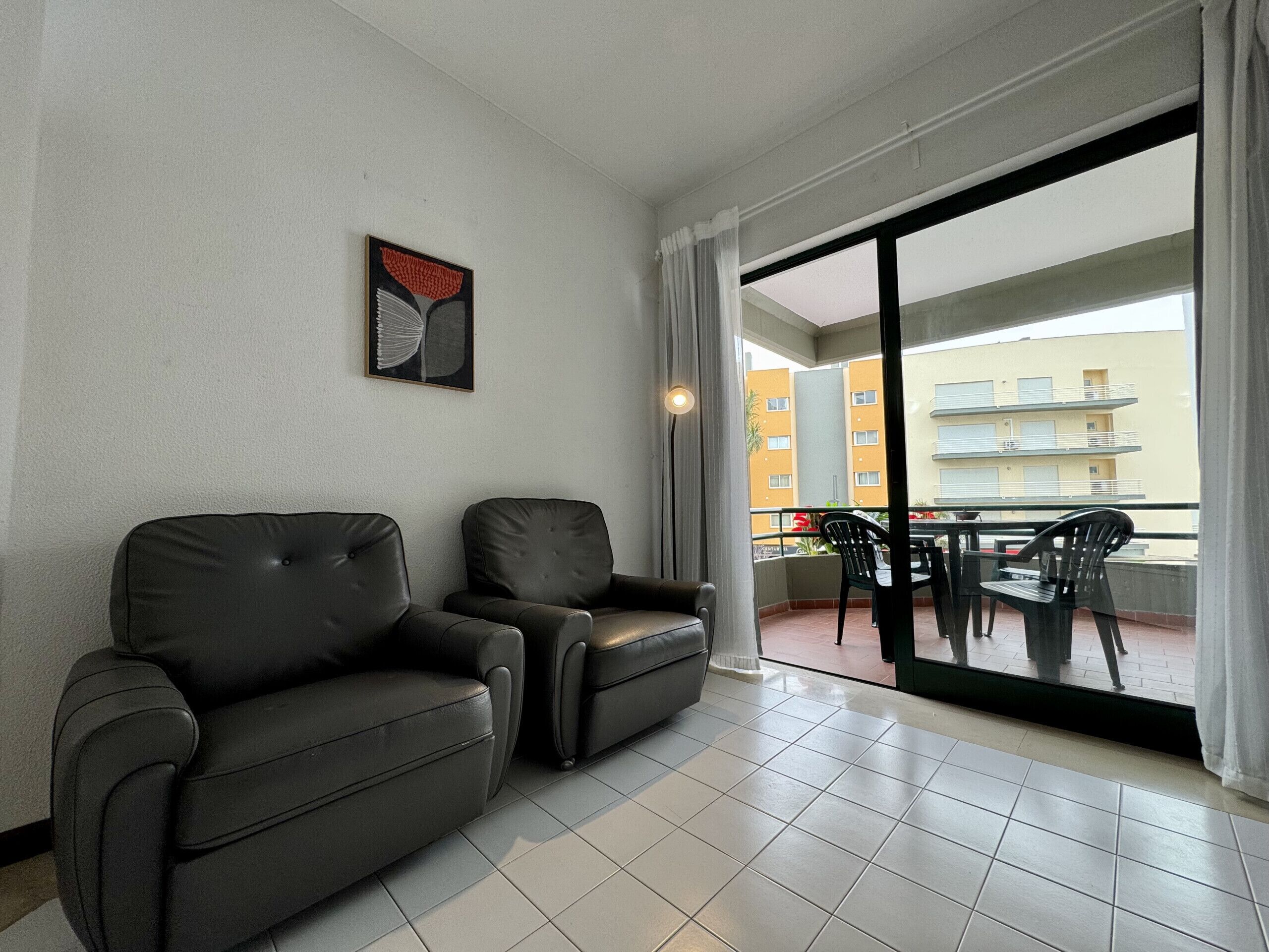 A modern living room with two leather sofas, a dining table with chairs, and a large sliding glass door leading to an outdoor balcony with a view of a yellow building.