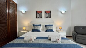 Ein Hotelzimmer mit einem Doppelbett, zwei Nachttischen, einem grauen Sessel und Kunstwerken an den Wänden.