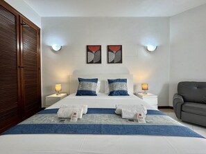 Ein Hotelzimmer mit einem Doppelbett, zwei Nachttischen, einem grauen Sessel und Kunstwerken an den Wänden.