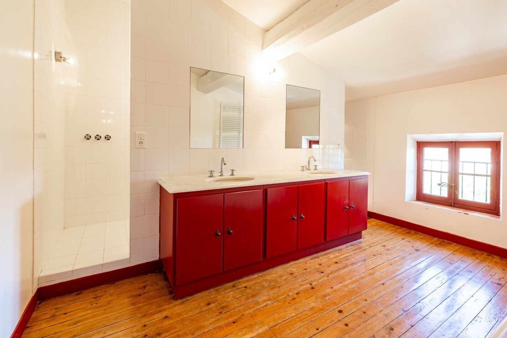 A bathroom with a wooden floor, a red vanity, a large mirror, and a window with a view.
