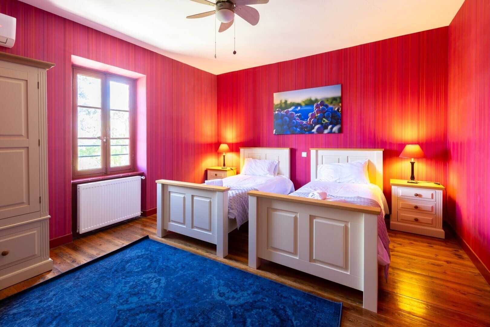 Une chambre avec deux lits, un ventilateur de plafond, une fenêtre et un tableau au mur.