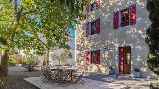 Ein Gebäude mit roten Fensterläden, einer Terrasse mit Tisch und Stühlen und einem Baum, der Schatten spendet.