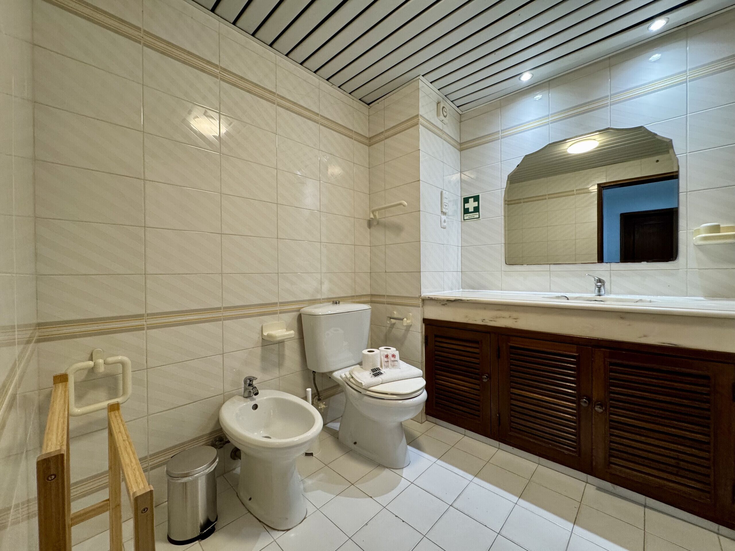 A bathroom with white tiles, a bidet, a toilet, and wooden cabinets.