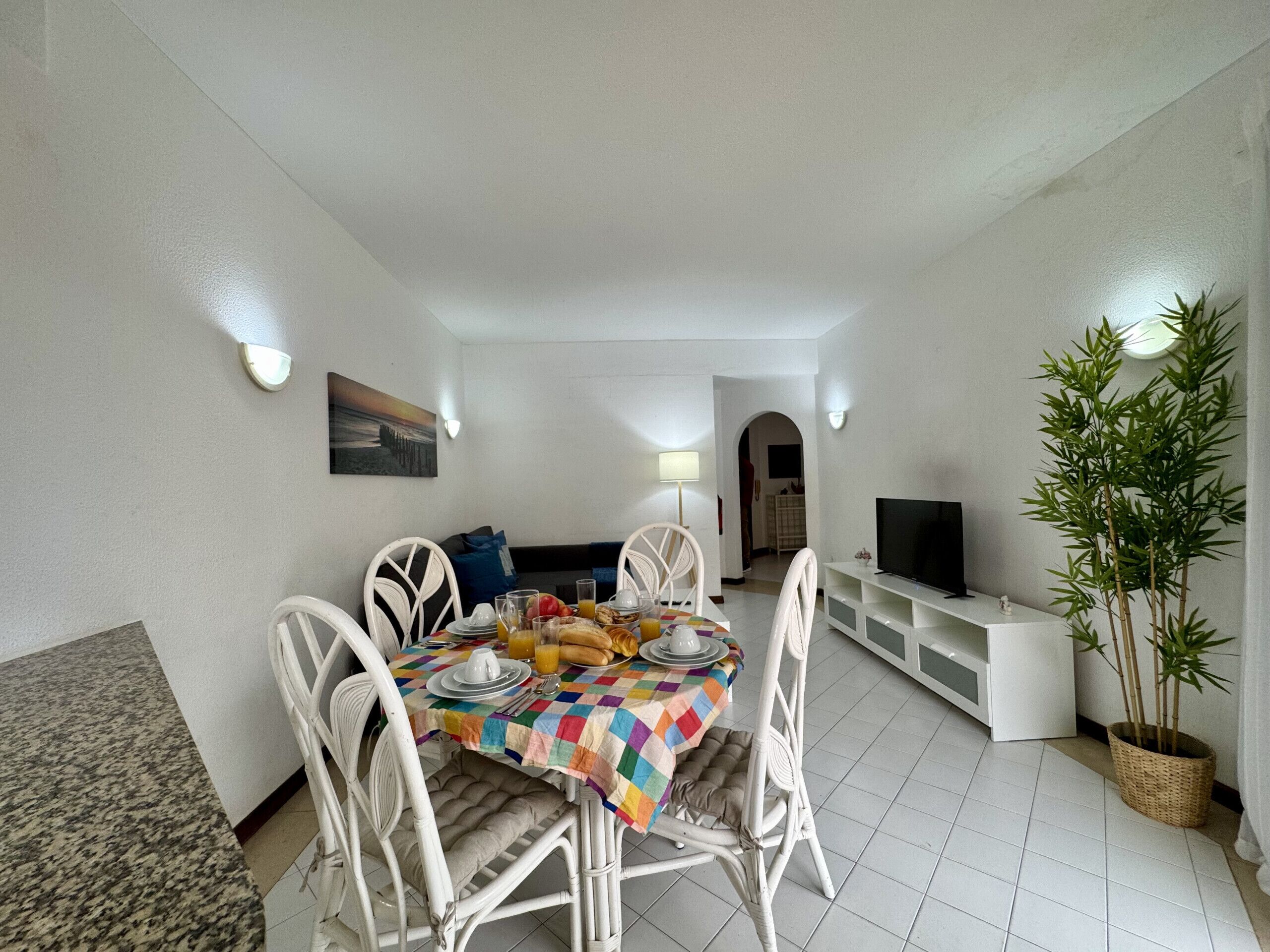 A dining area with a table set for breakfast, white chairs, a television, and a potted plant.