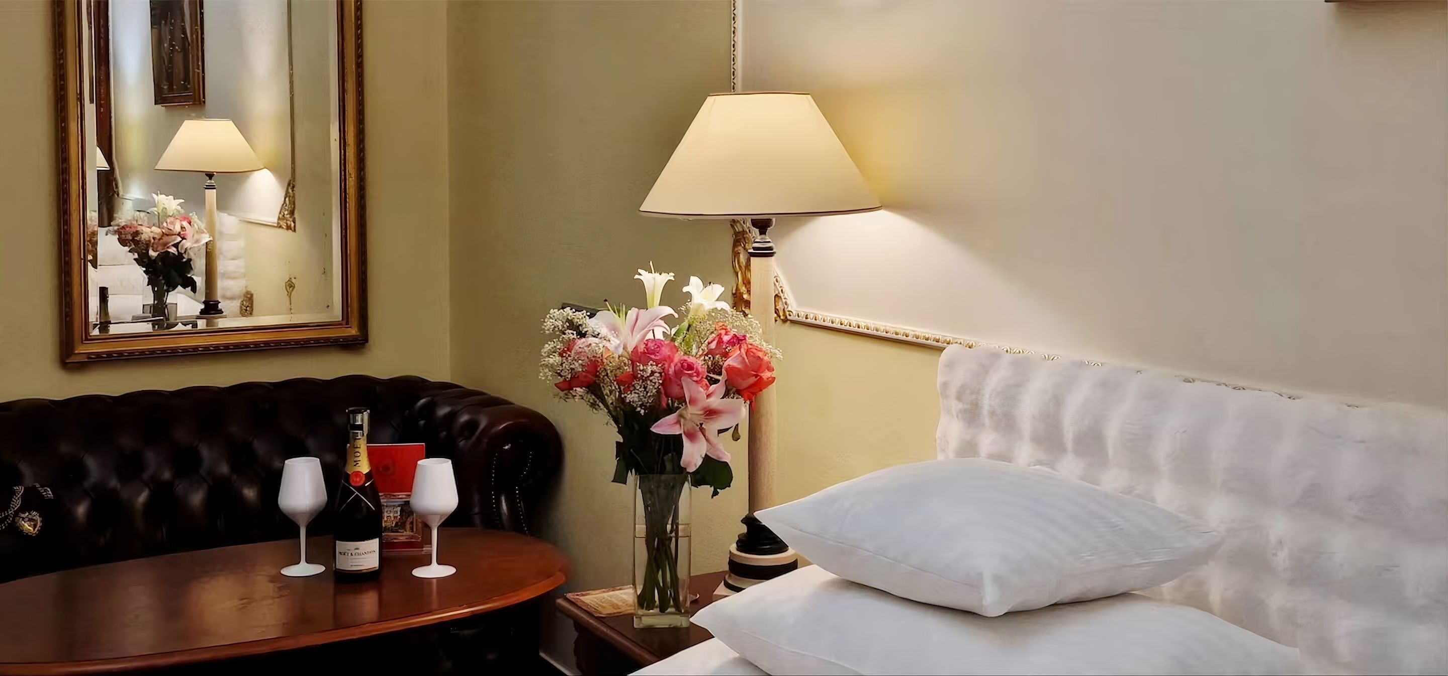 Quarto de hotel com um sofá de couro escuro, uma mesa redonda de madeira com uma garrafa de champanhe, duas taças de vinho, um vaso de flores, um abajur e uma cama com almofadas brancas.