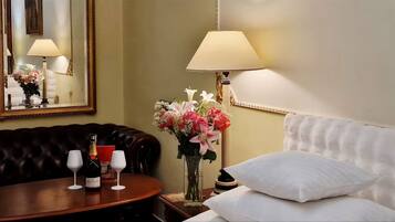 Quarto de hotel com um sofá de couro escuro, uma mesa redonda de madeira com uma garrafa de champanhe, duas taças de vinho, um vaso de flores, um abajur e uma cama com almofadas brancas.