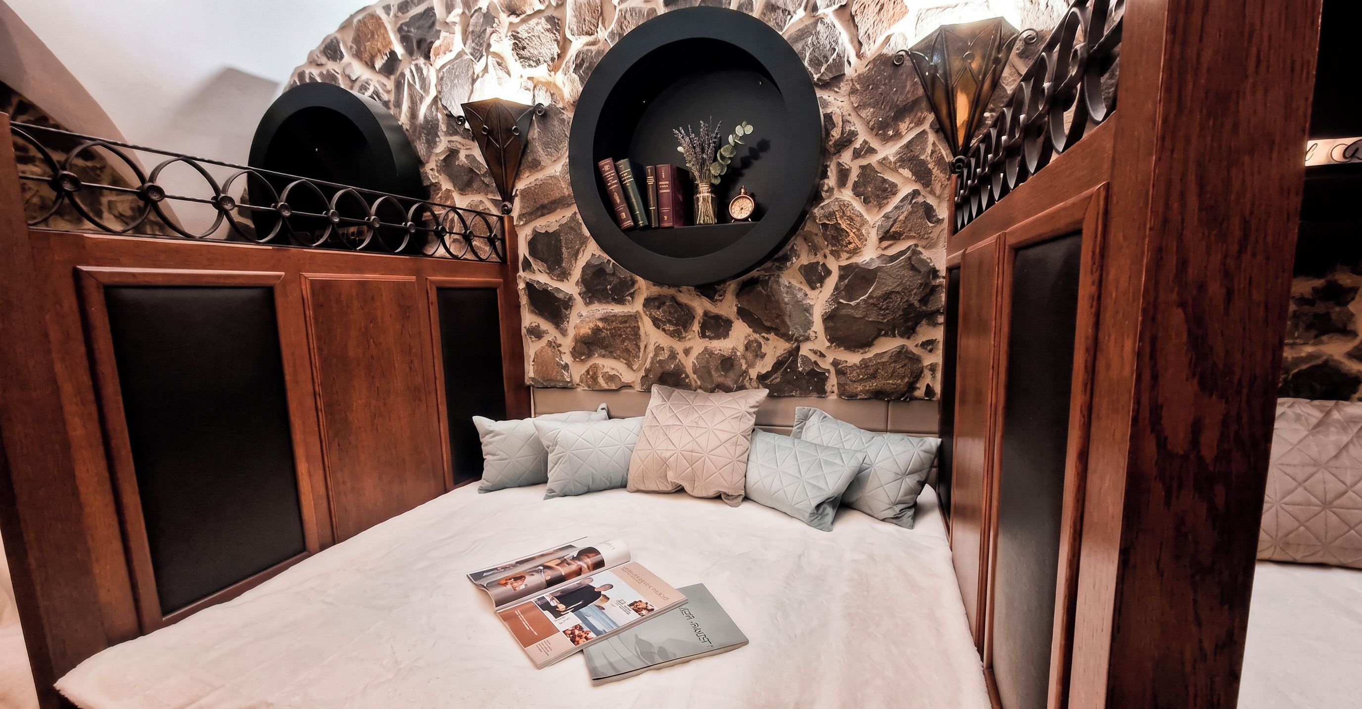 Une chambre avec un mur en pierre, un lit avec un magazine, et une étagère avec des livres et une boussole.