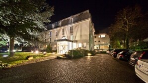 A building with a paved driveway, a covered entrance, and parked cars. - Select Hotel Silence Garden (Cologne)