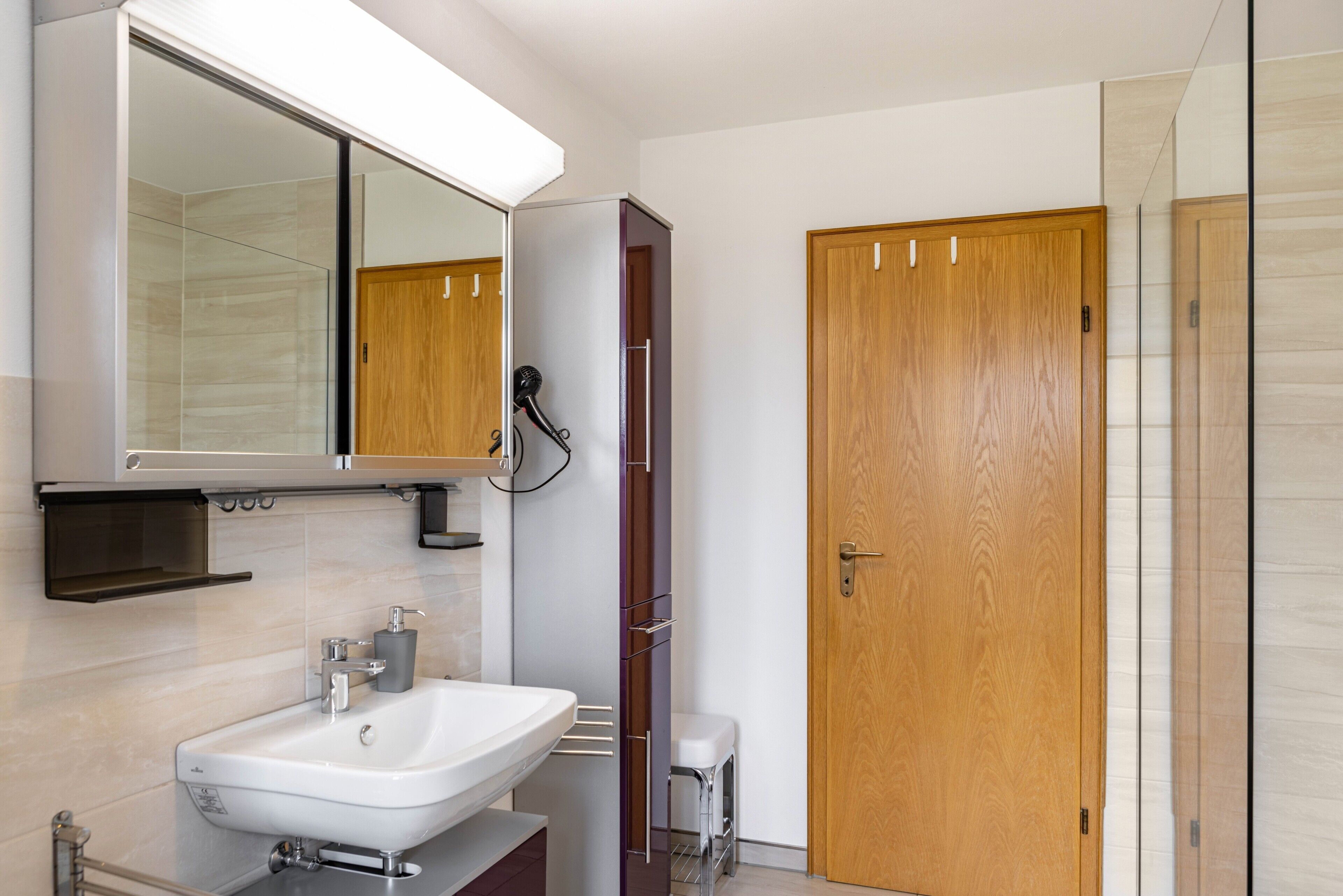 Une salle de bain moderne avec un lavabo blanc, un miroir, un sèche-cheveux et une porte en bois.