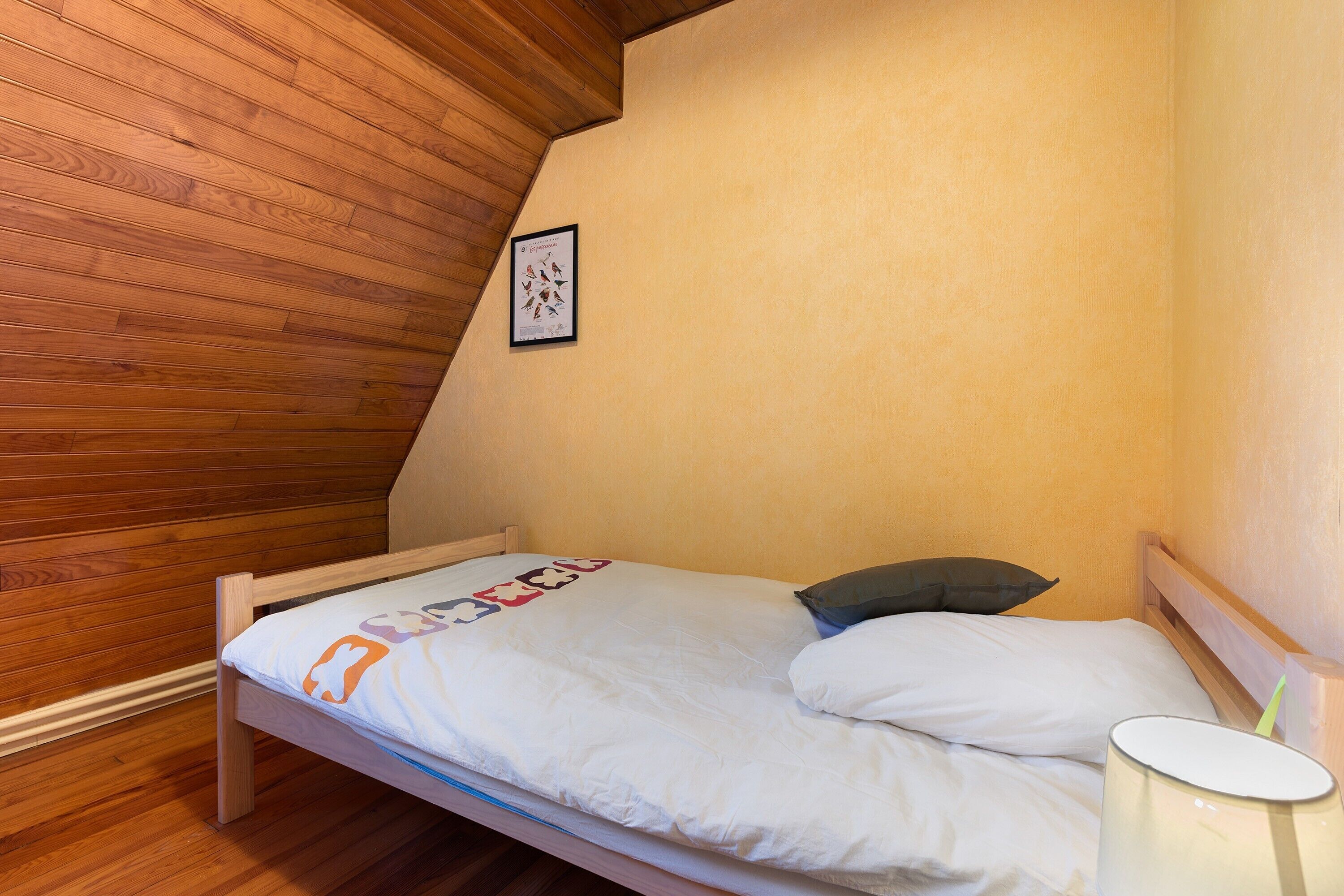 Una cama individual con colchón blanco y almohada, cabecera de madera, un cuadro enmarcado en la pared y una lámpara de noche.