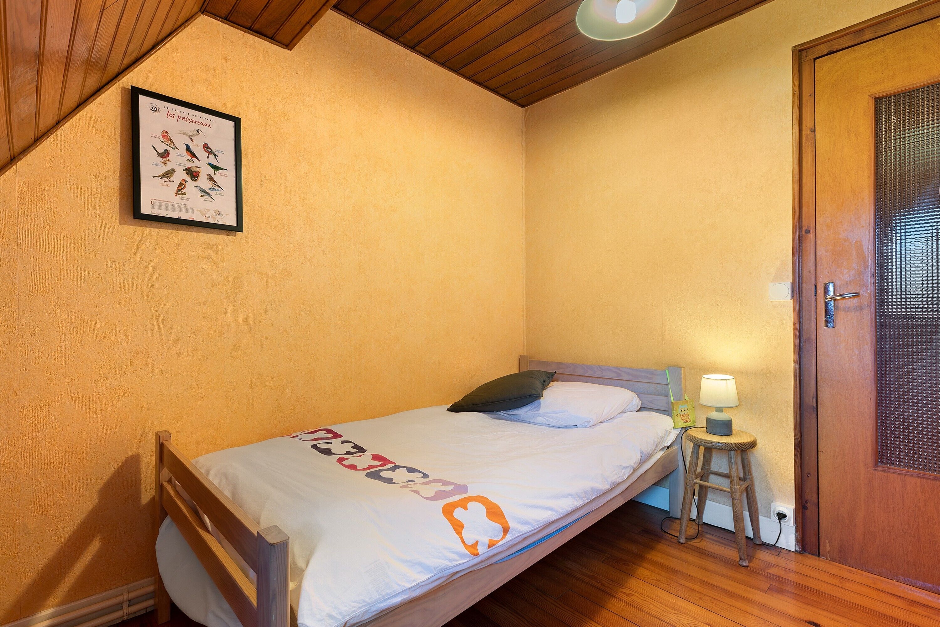 A single bed with a white mattress and a blanket, a wooden headboard, a small stool with a lamp, and a door with a metal screen.