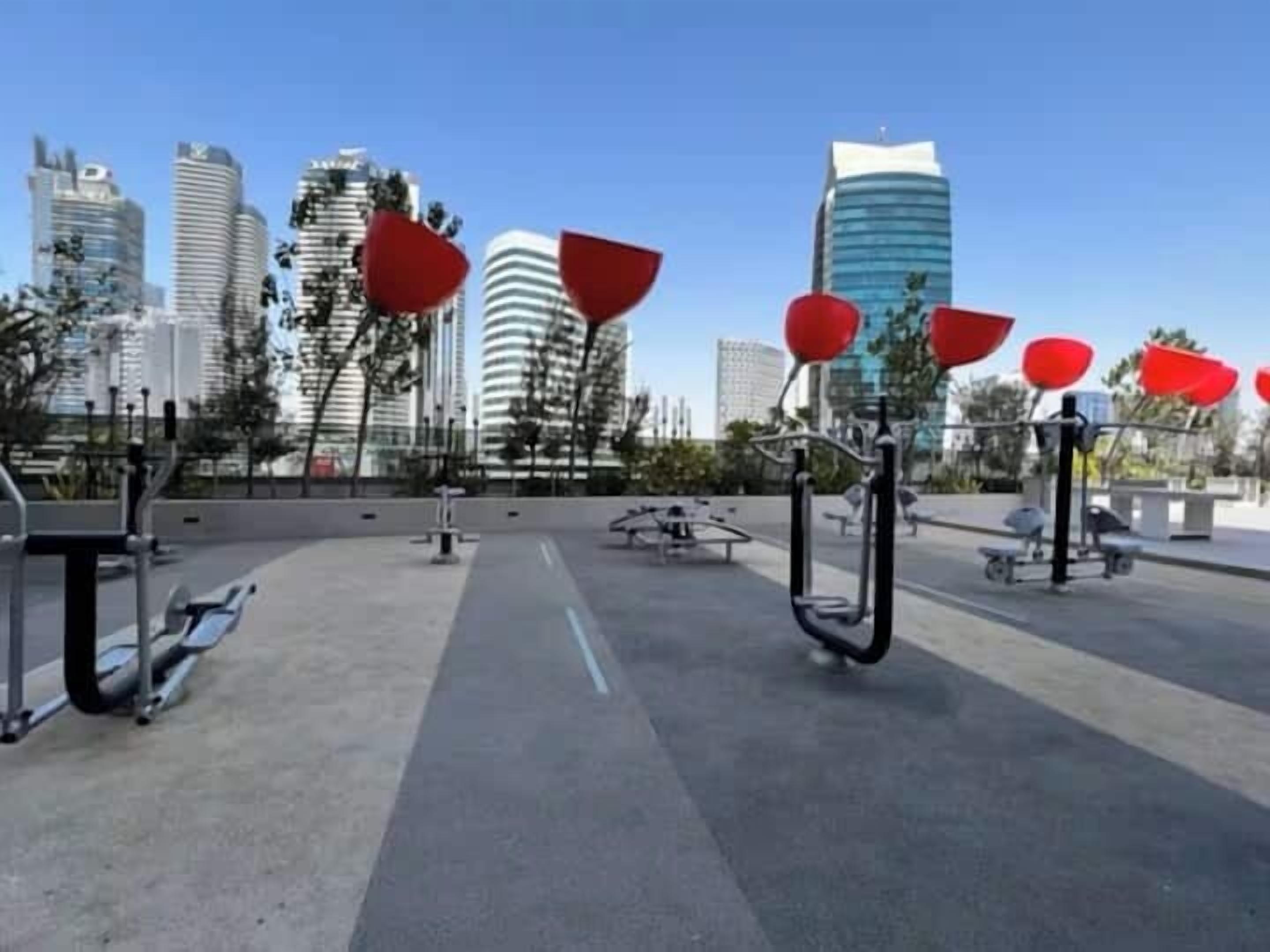 An outdoor exercise area with stationary bikes and red exercise equipment, surrounded by modern buildings.
