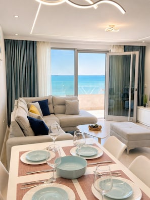 A modern living room with a sofa, dining table set for two, and a view of the sea through large windows.