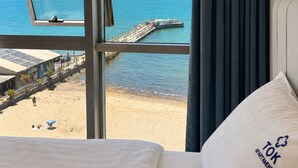 Hotelkamer met een groot raam met uitzicht op een strand, een bed met een kussen waarop 'TOK APARTMENTS' staat, en een zeegezicht met een pier.