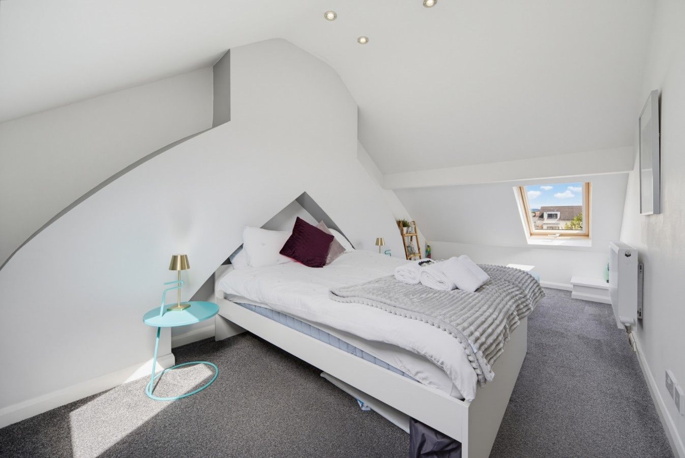Une chambre moderne avec un plafond en pente, un lit avec une literie blanche et un oreiller violet, une petite table ronde avec une lampe, et une fenêtre donnant sur l’extérieur.