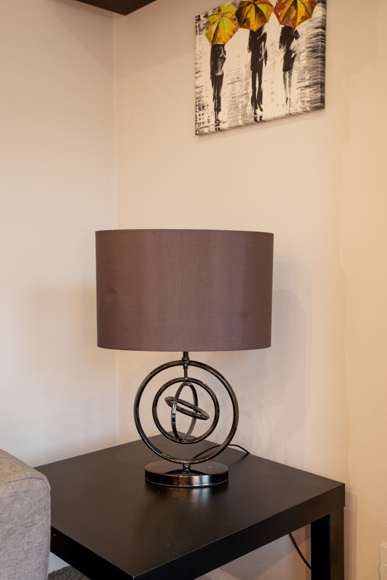 Une lampe de table moderne, dotée d’un pied circulaire et d’un abat-jour rectangulaire, posée sur une table sombre, devant un mur uni.