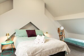 A bedroom with a pitched ceiling, a bed with a green and maroon pillow, a white blanket, and a teal bench. There are two bedside tables with lamps and a wooden ladder. - Central Cardiff 2-Bed Flat Near Stadium (Cardiff)