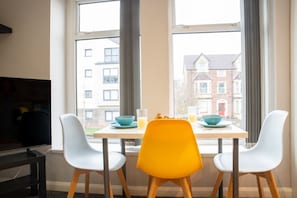 A dining area with a table set for two, two chairs, a TV, and a window with a view of buildings. - Central Cardiff 2-Bed Flat Near Stadium (Cardiff)