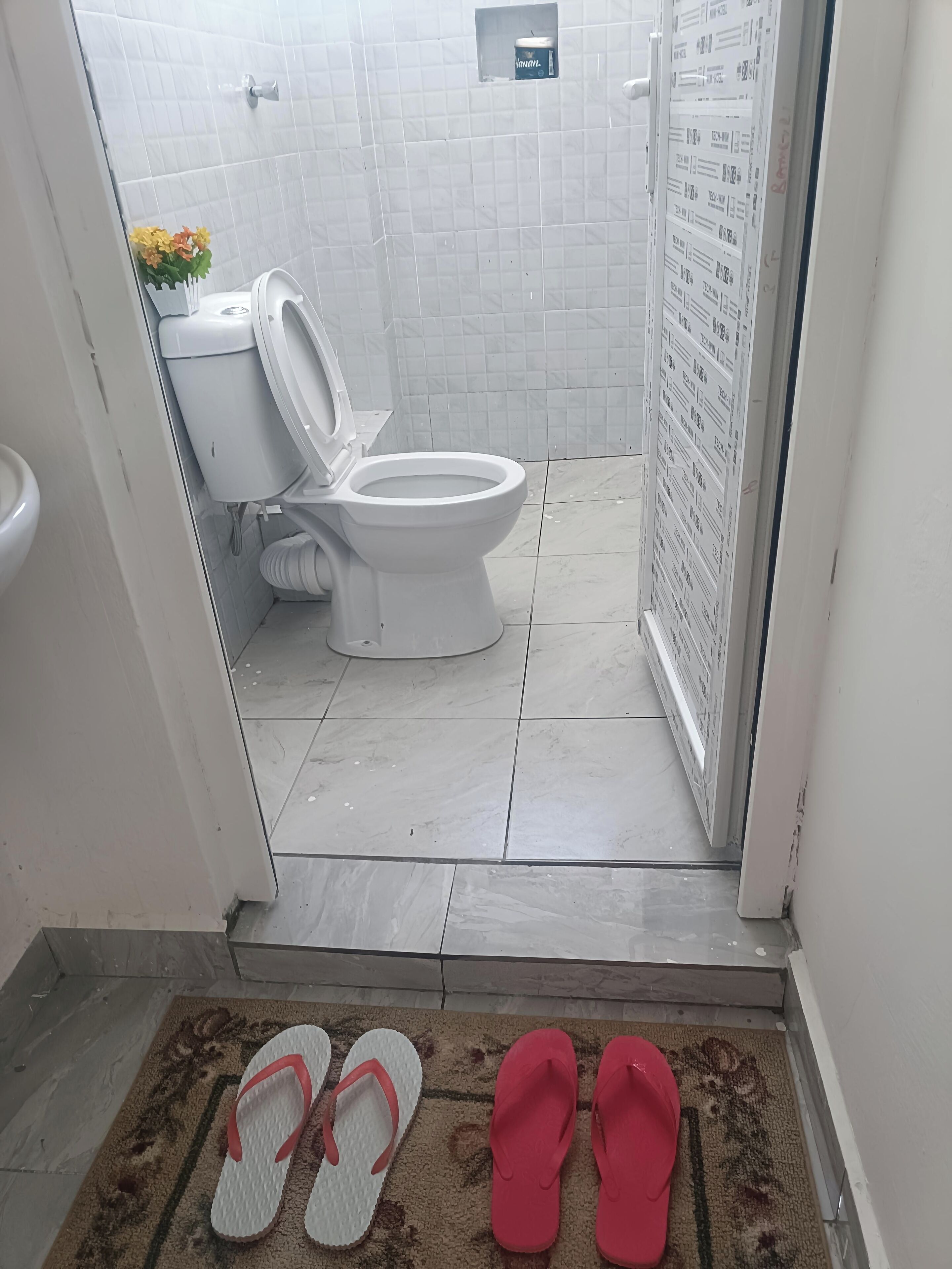 Ein Badezimmer mit Toilette, Duschbereich und einem Paar Badeschlappen auf einer Fußmatte.