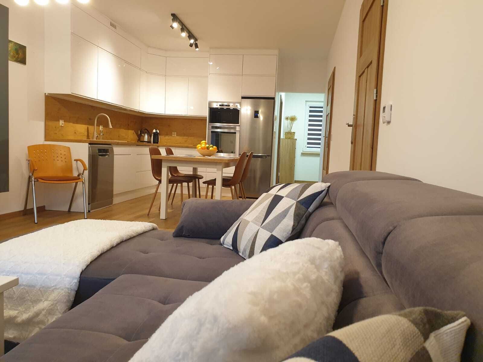 A modern living room with a grey sofa, a white fluffy throw, and a checkered pillow. A wooden dining table with chairs is in the background, and a kitchen with white cabinets and a stainless steel refrigerator is visible.
