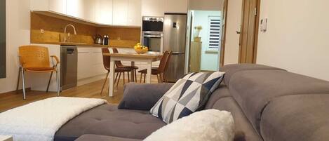 A modern living room with a grey sofa, a white fluffy throw, and a checkered pillow. A wooden dining table with chairs is in the background, and a kitchen with white cabinets and a stainless steel refrigerator is visible.