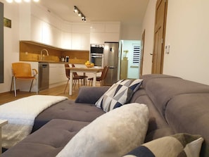 Una sala de estar moderna con un sofá gris, un plaid blanco y mullido, y una almohada a cuadros. En el fondo, una mesa de comedor de madera con sillas, y se ve una cocina con armarios blancos y un refrigerador de acero inoxidable.