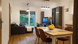 En moderne stue med sofa, spisebord med stole, fjernsyn og et vindue med udsigt til grønne omgivelser.