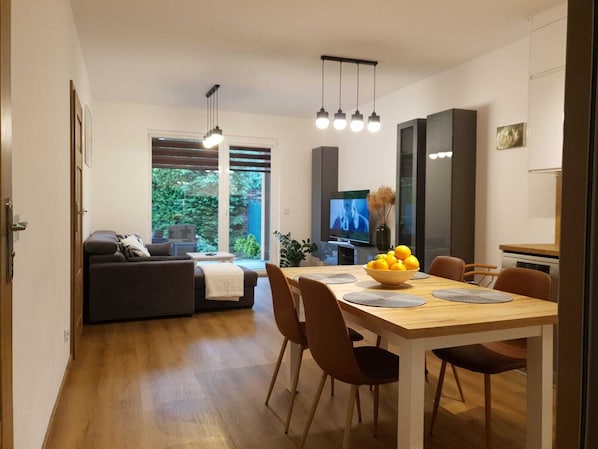Una sala de estar moderna con un sofá, una mesa de comedor con sillas, un televisor y una ventana que da a zonas verdes.