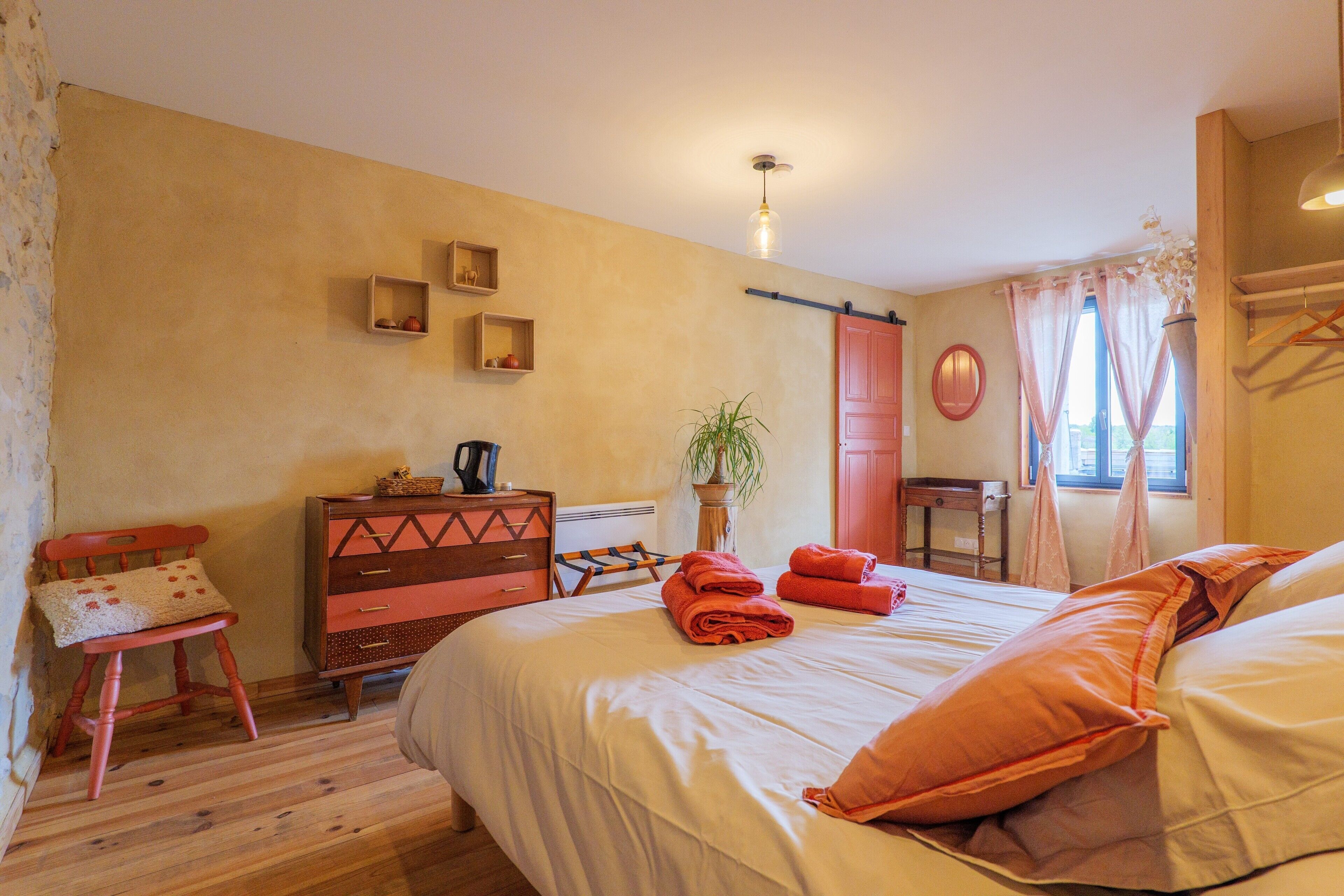 Un dormitorio con cama, cómoda, silla, planta y ventana con cortinas.