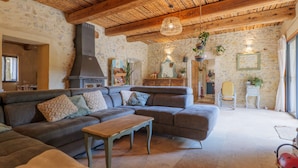 Un salon chaleureux avec un mur en pierre, des poutres en bois au plafond, un grand canapé, une petite table et un luminaire suspendu.